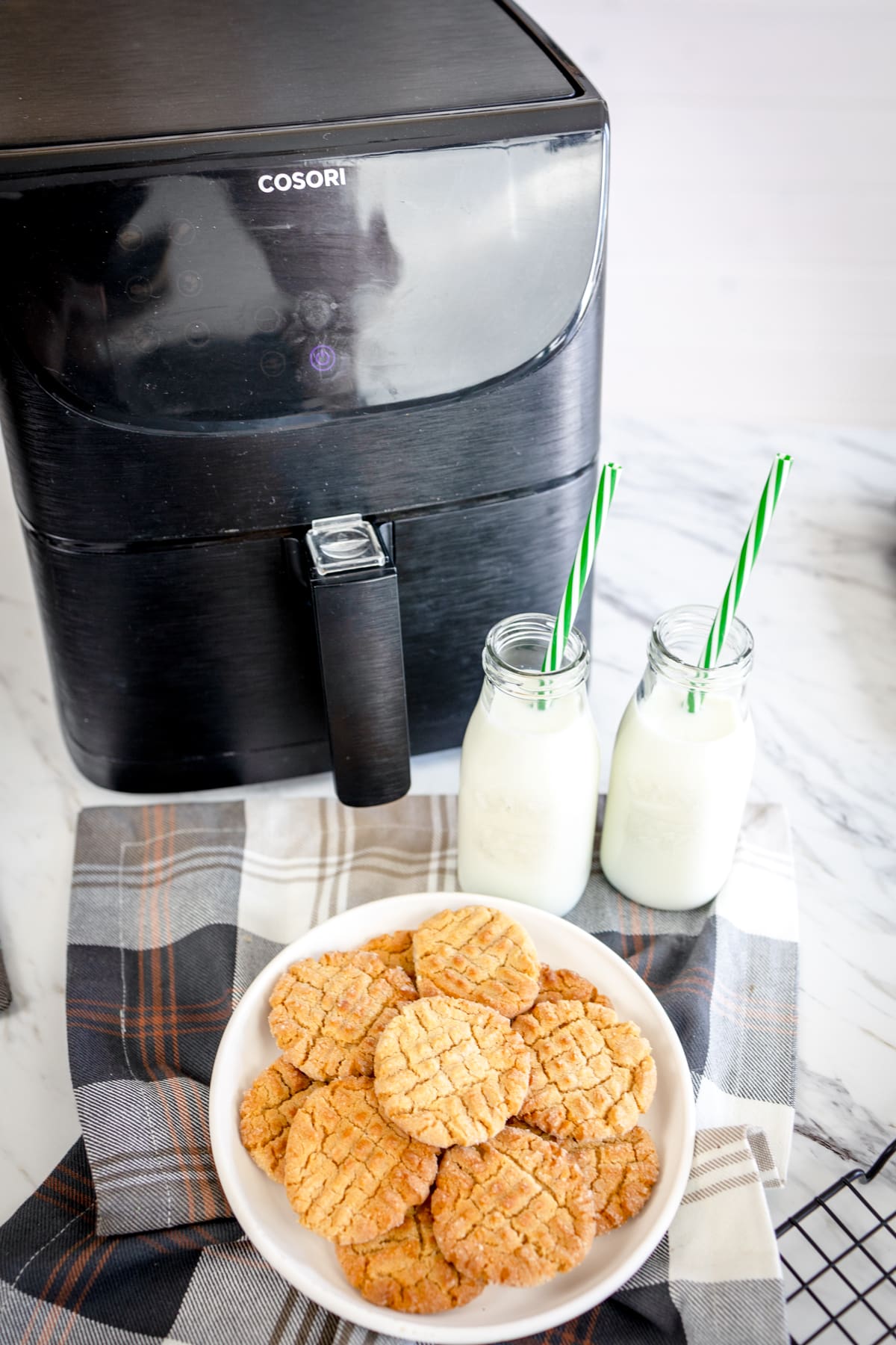 A plate of peanut butter cookies on a table next to an air fryer and a glass of milk.