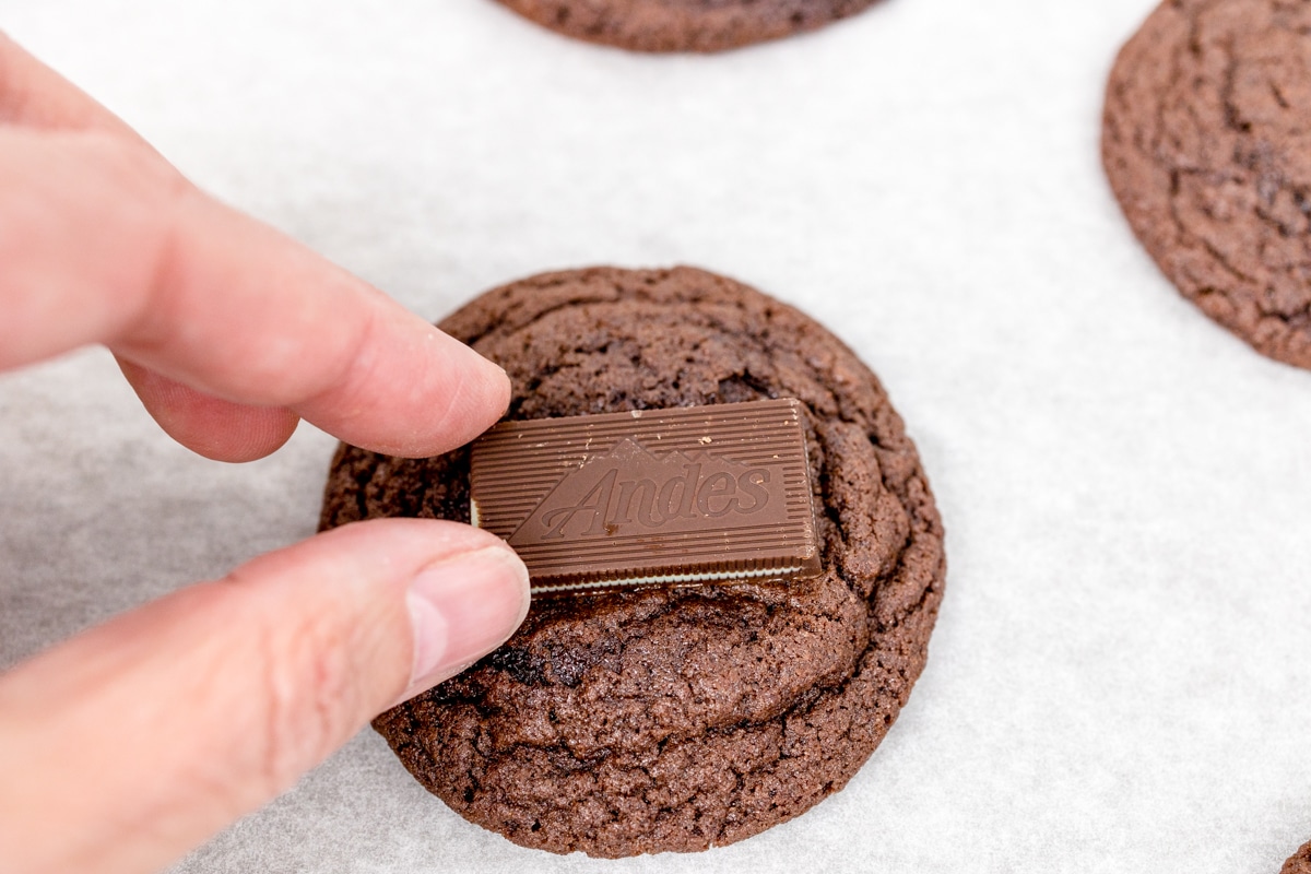 Close up of a chocolate mint cookie with a andes mint chocolate being placed on top to melt. 