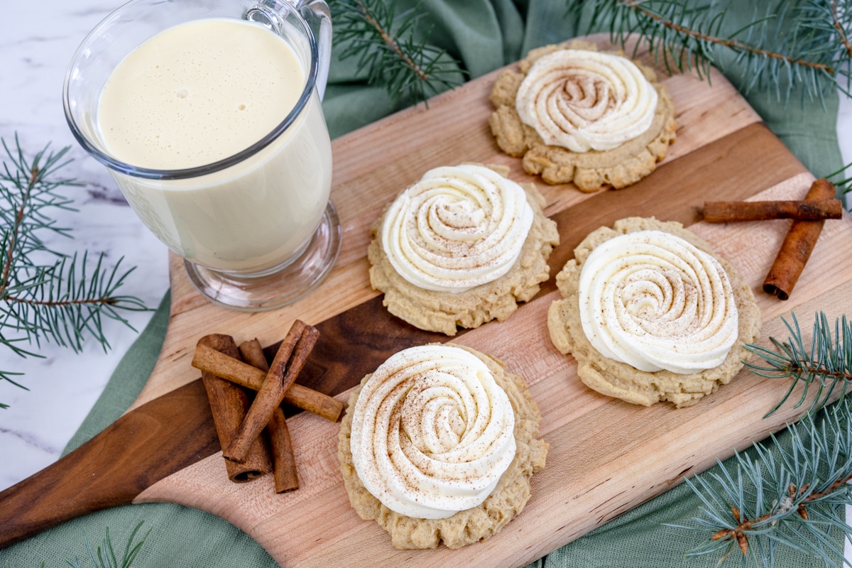 Eggnog cookies with frosting on a tray with cinnamon sticks on it too and a glass of eggnog.