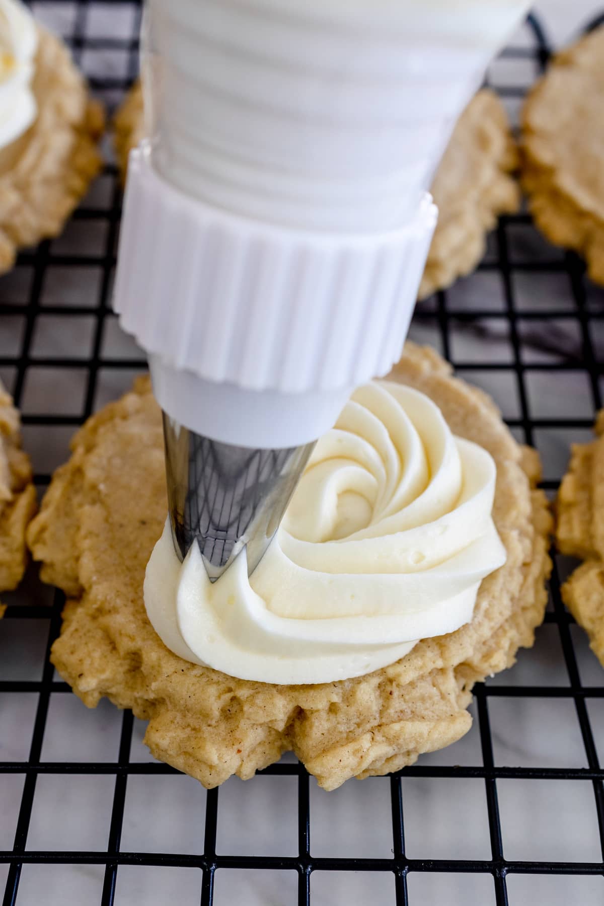 Close up of an eggnog cookie on a wire rack being frosted with a piping bag and thick cream frosting.  