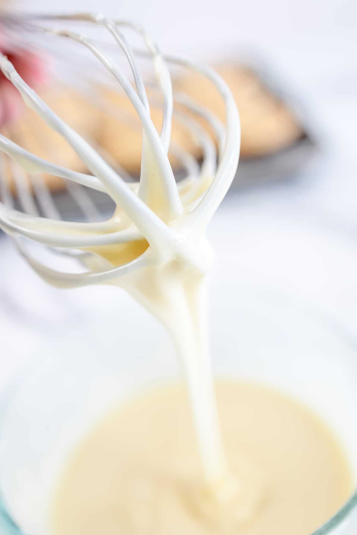 Close up of a whisk coated in white glaze that is dripping off into a glass bowl below.