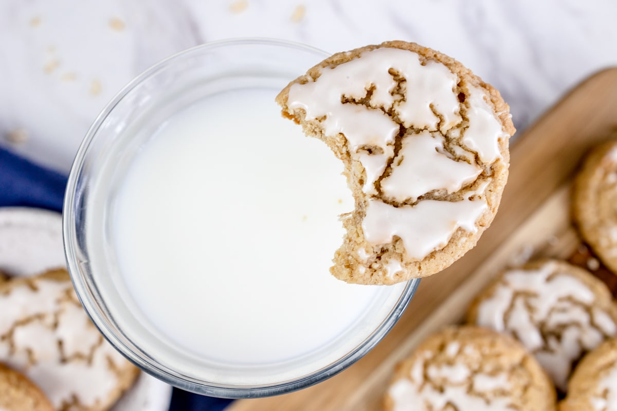 Close up of an iced oatmeal cookie with a bite taken out of it, balancing on the top rim of a glass full of milk.