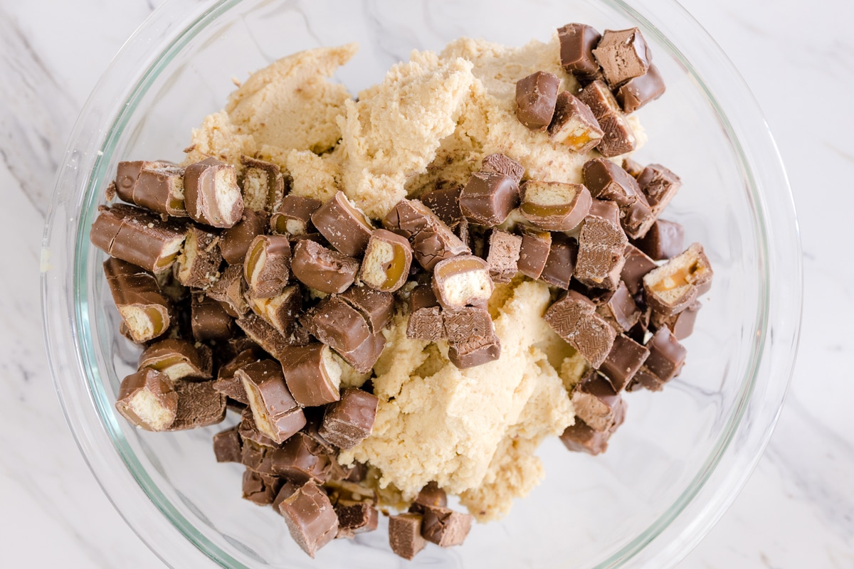 Top view of a glass bowl with candy cookie dough and chopped chocolate bars being mixed by a silver spoon. 