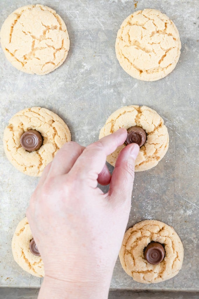 Top view of peanut butter cookies on a baking tray being topped with a Rolo candy.