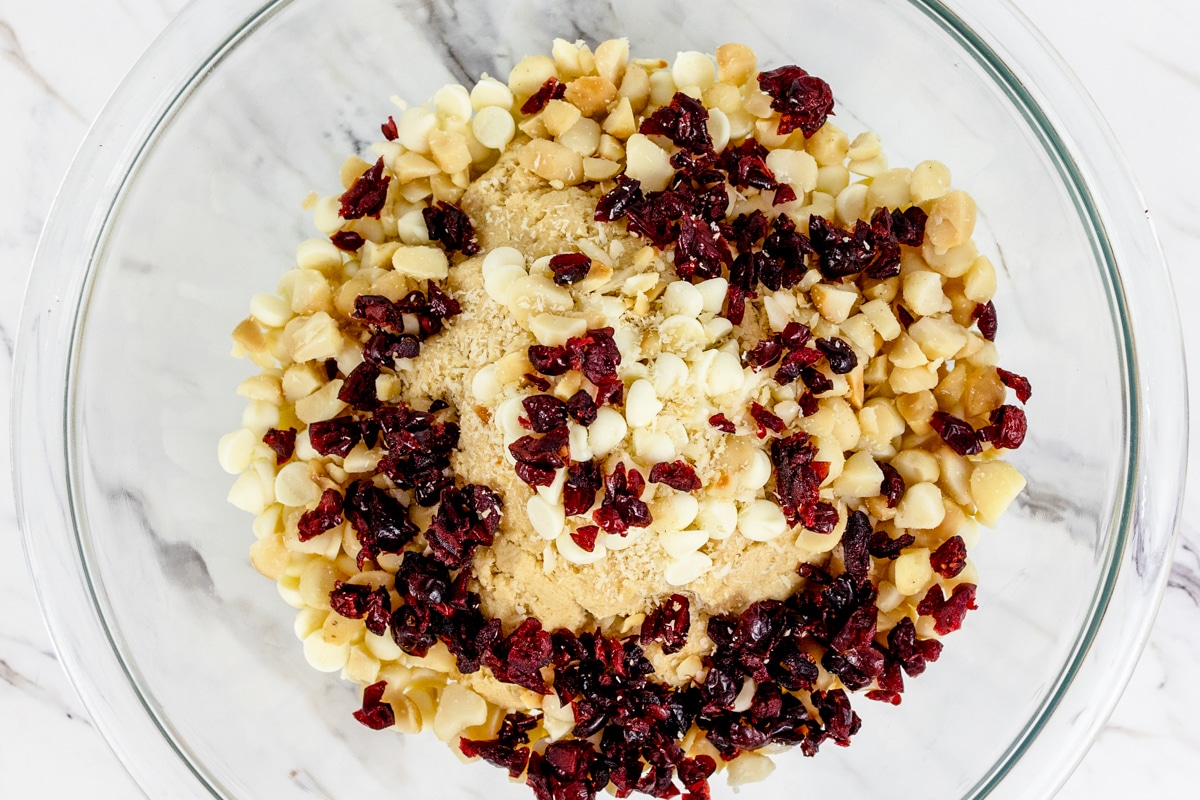 Top view of glass bowl with cookie dough topped with white chocolate chips, macadamia nuts and cranberries.