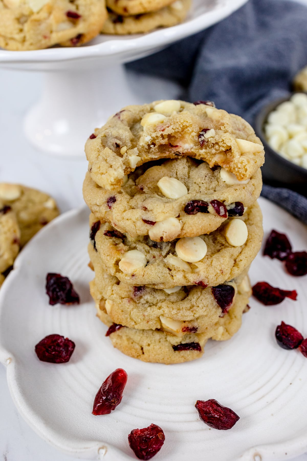 White chooclate macadamia nut cookies stacked on a white plate with canberries dotted around them.