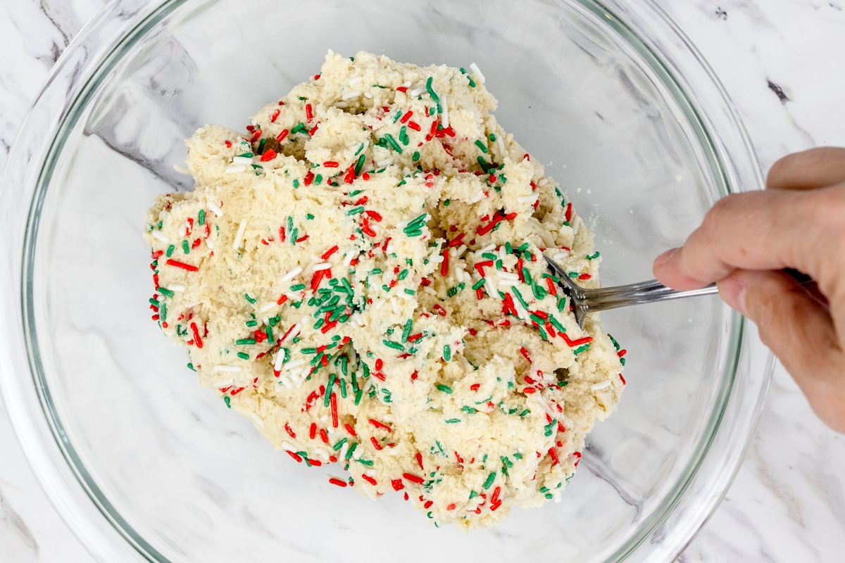 Top view of a glass bowl with sugar cookie mix in it with red and green sprinkles being stirred into it with a spoon.