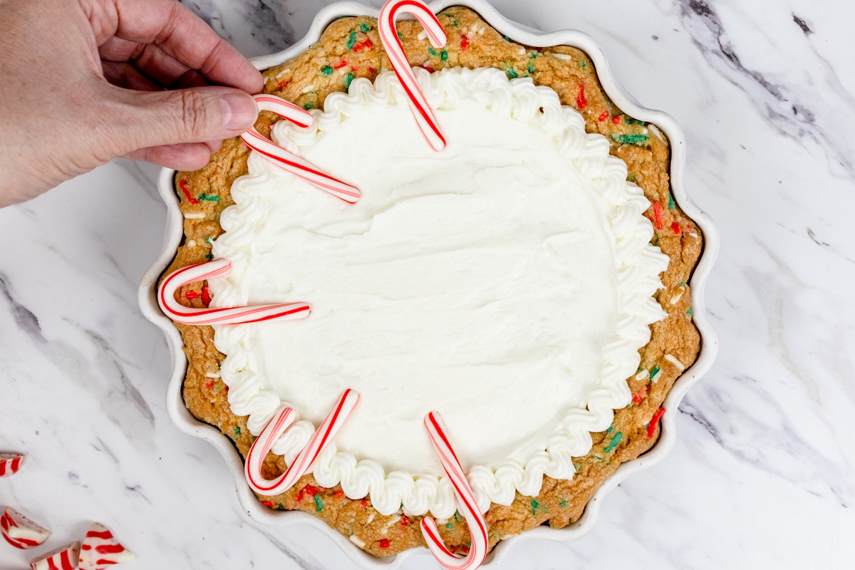 Top view of festive cookie pie with frosting on it and candy canes being placed evenly around the edges.