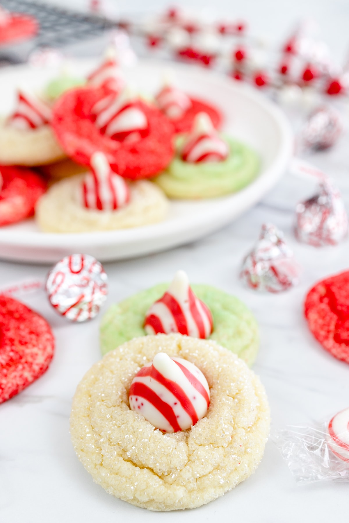 Two Peppermint Candy Cane Kisses Cookies on a worktop with a plate piled with the same cookies in the background.