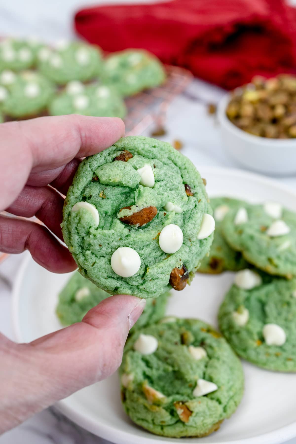 Close up of a pistachio and white chocolate cookie being held up by a hand, with more cookies on a plate in the background.