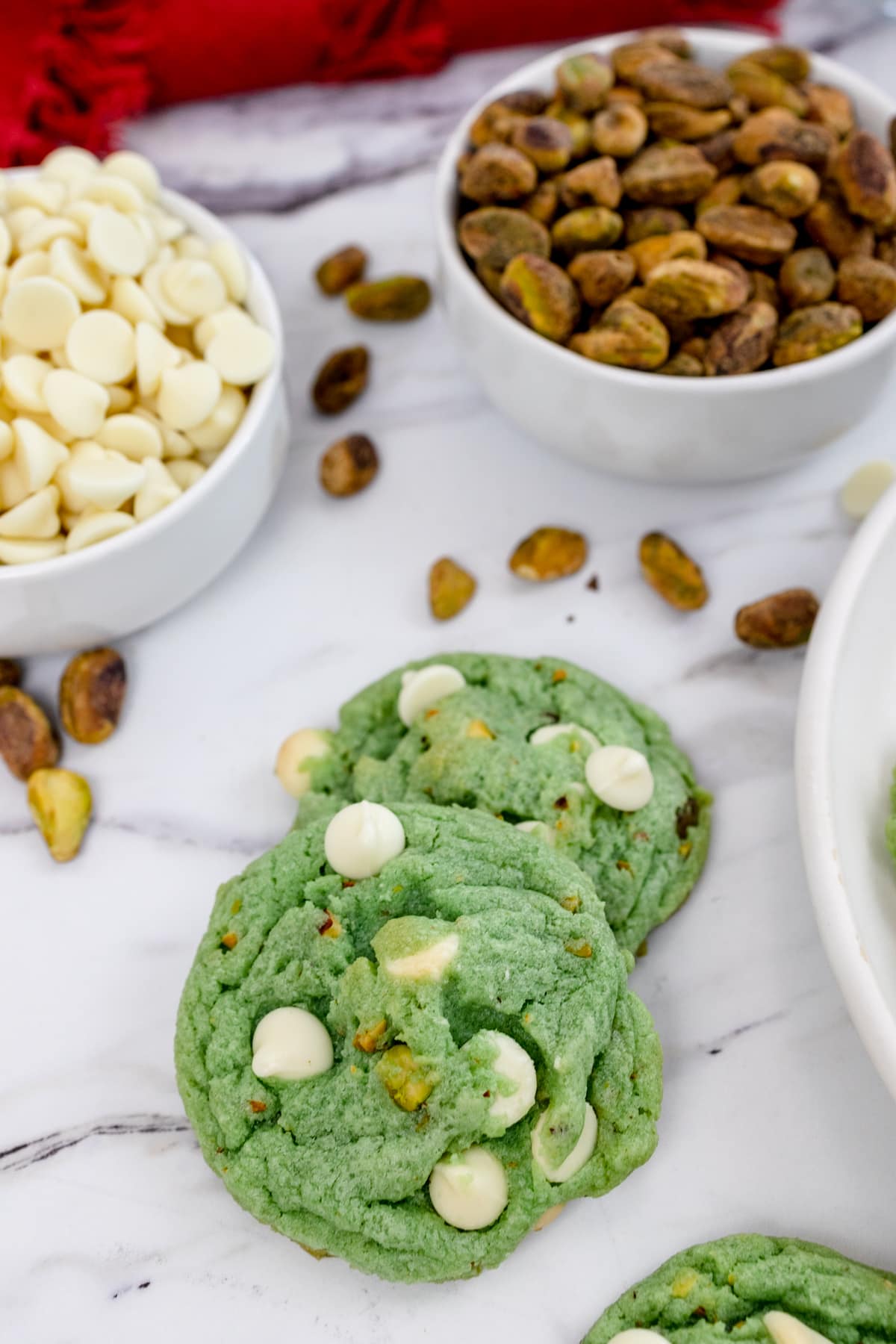 Close up of pistachio cookies on a table with small bowls of white chocolate chips and pistachio nuts next to them