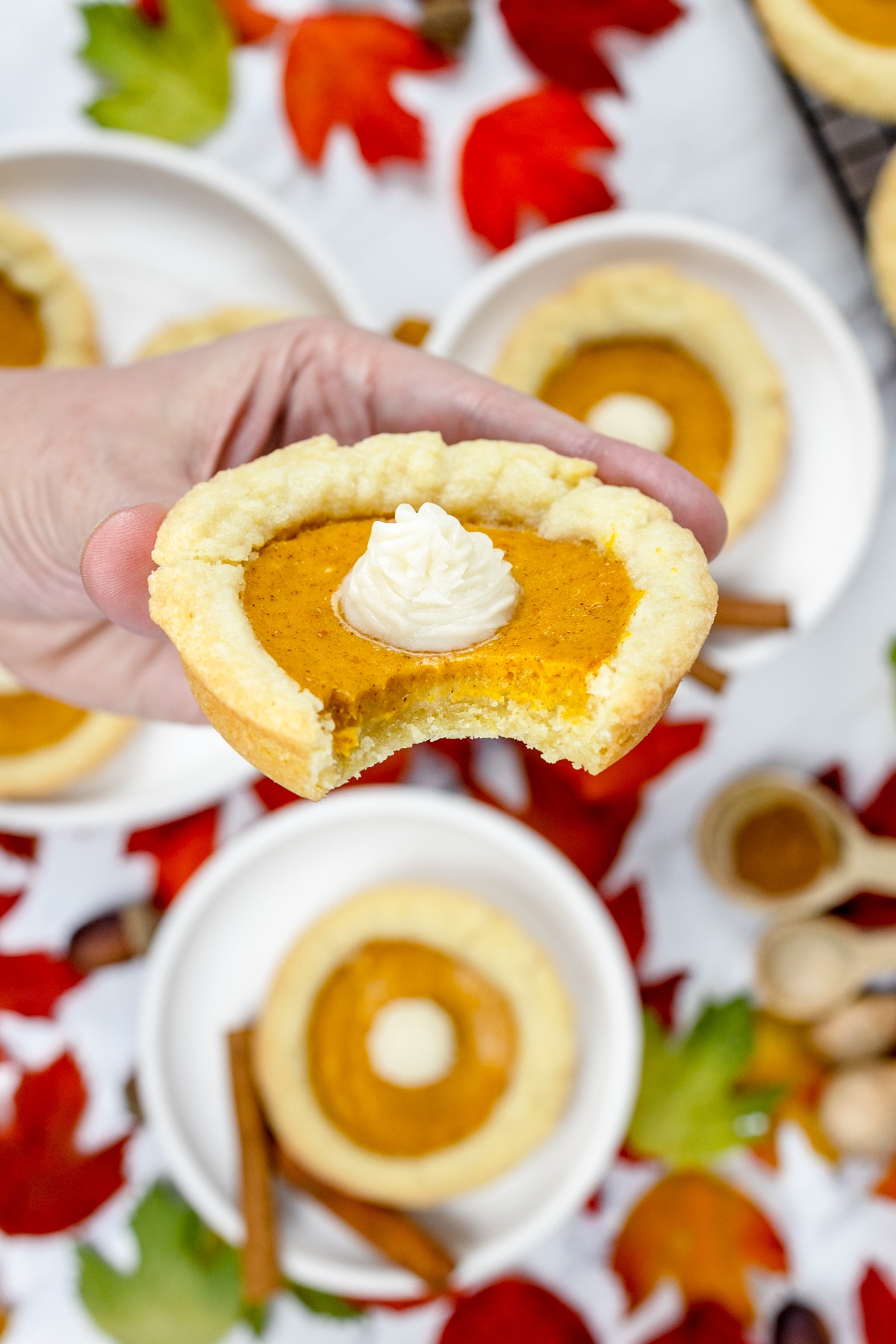 Close up of a mini pumpkin pie cookie with a dollop of frosting in the centre, being held up by a hand with more cookies in the background.