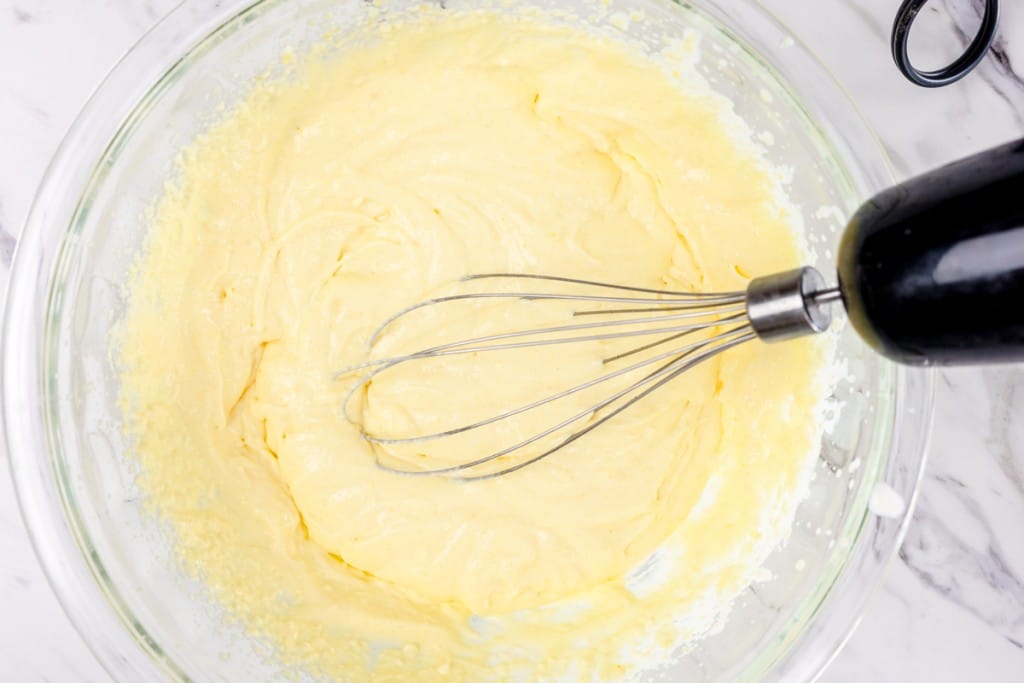 Top view of a glass mixing bowl with a yellow pudding mixture being mixed with a whisk.