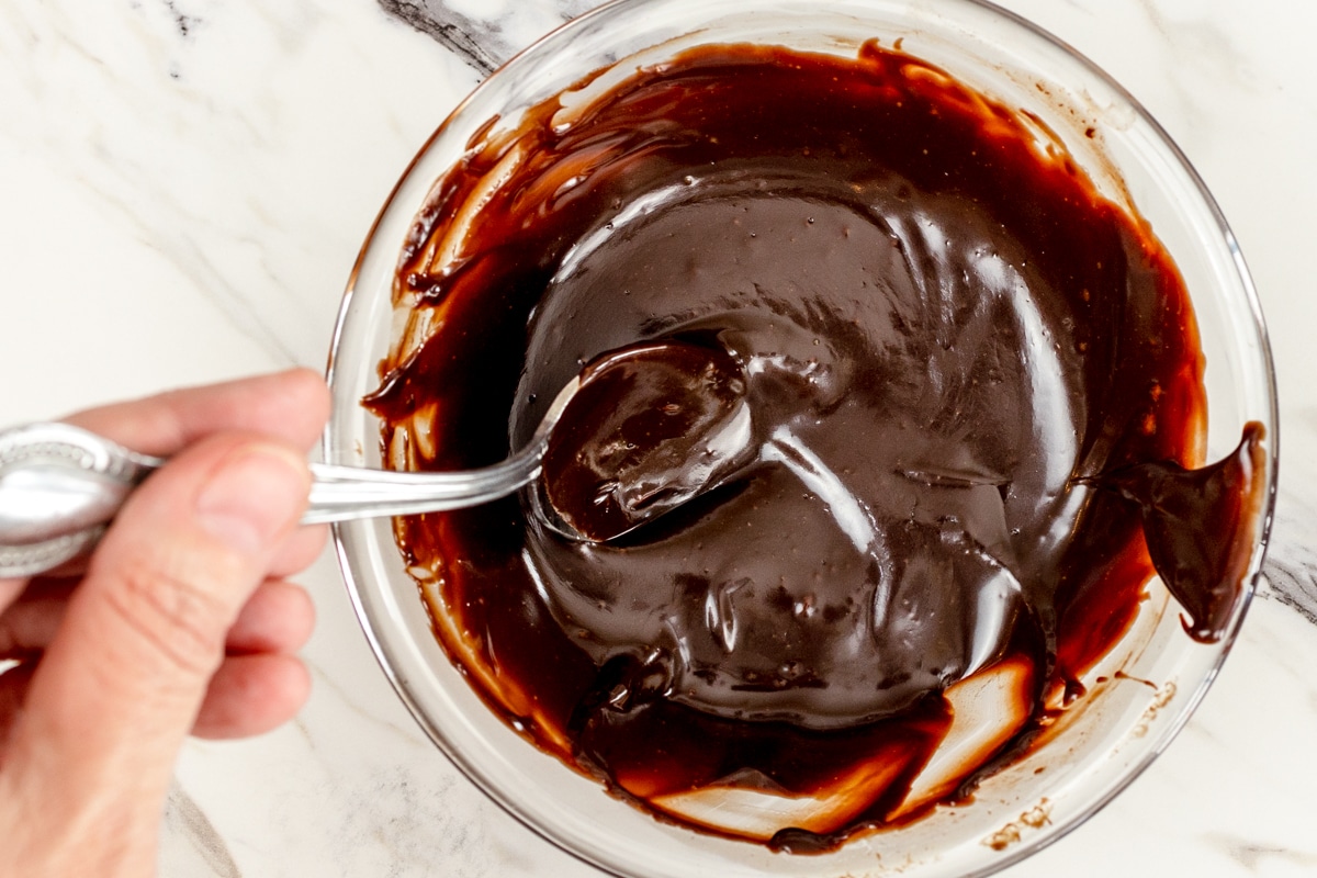 Top view of a glass mixing bowl with chocolate ganache in it being stirred with a spoon. 
