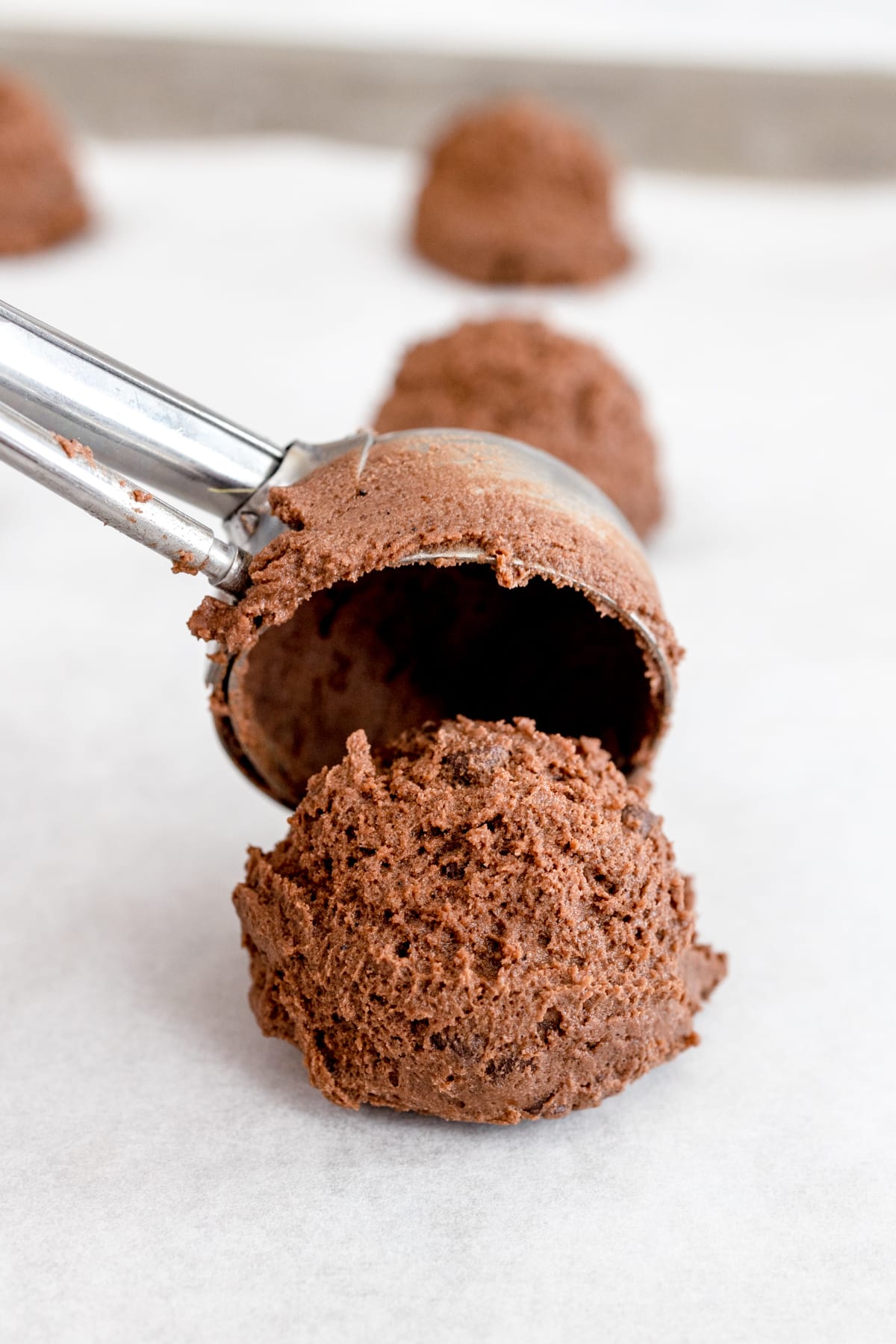 Close up of a chooclate cookie dough ball being placed on a baking sheet with a cookie scoop.