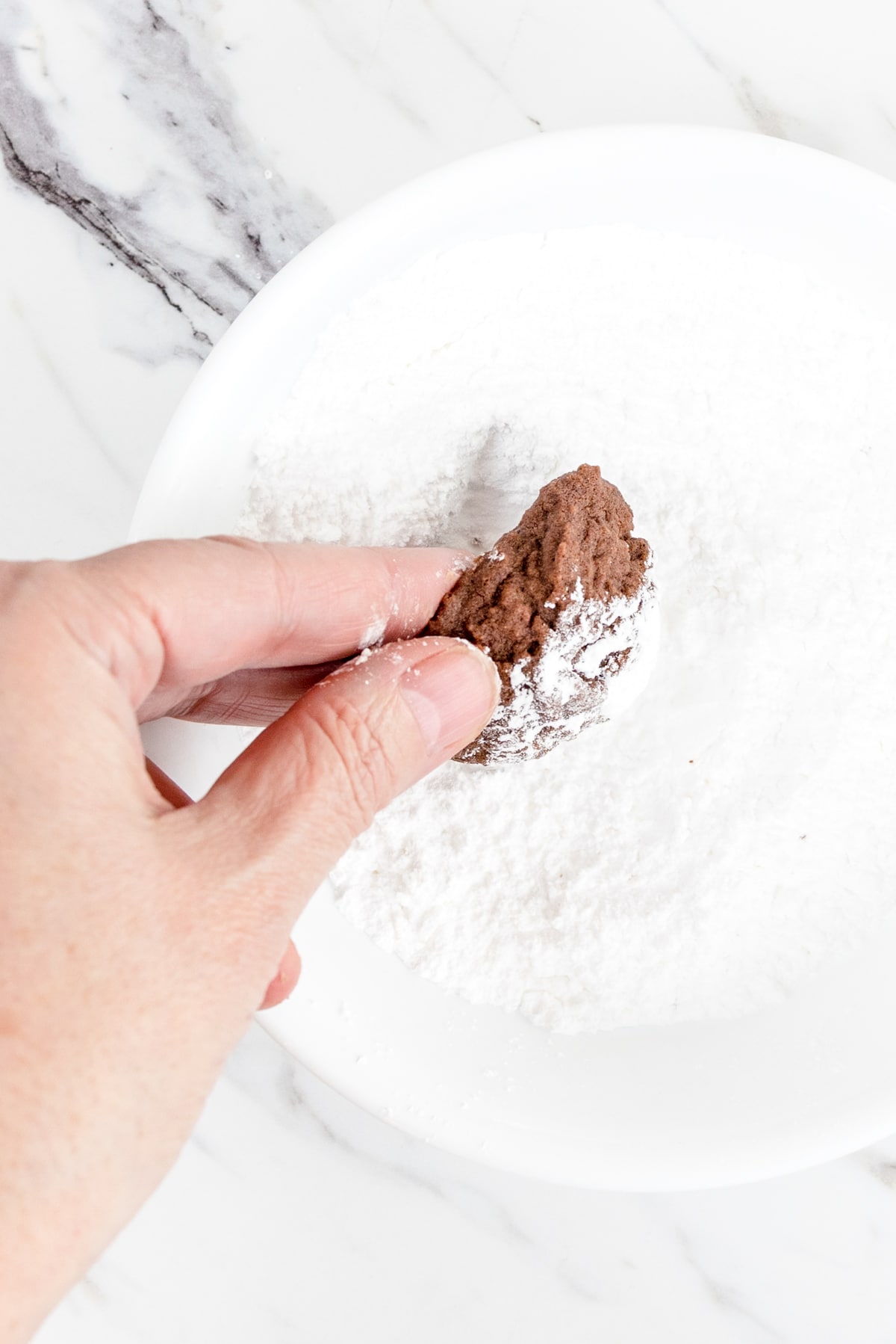 Top view of a chocolate cookie being coated in powdered sugar in a small bowl. 