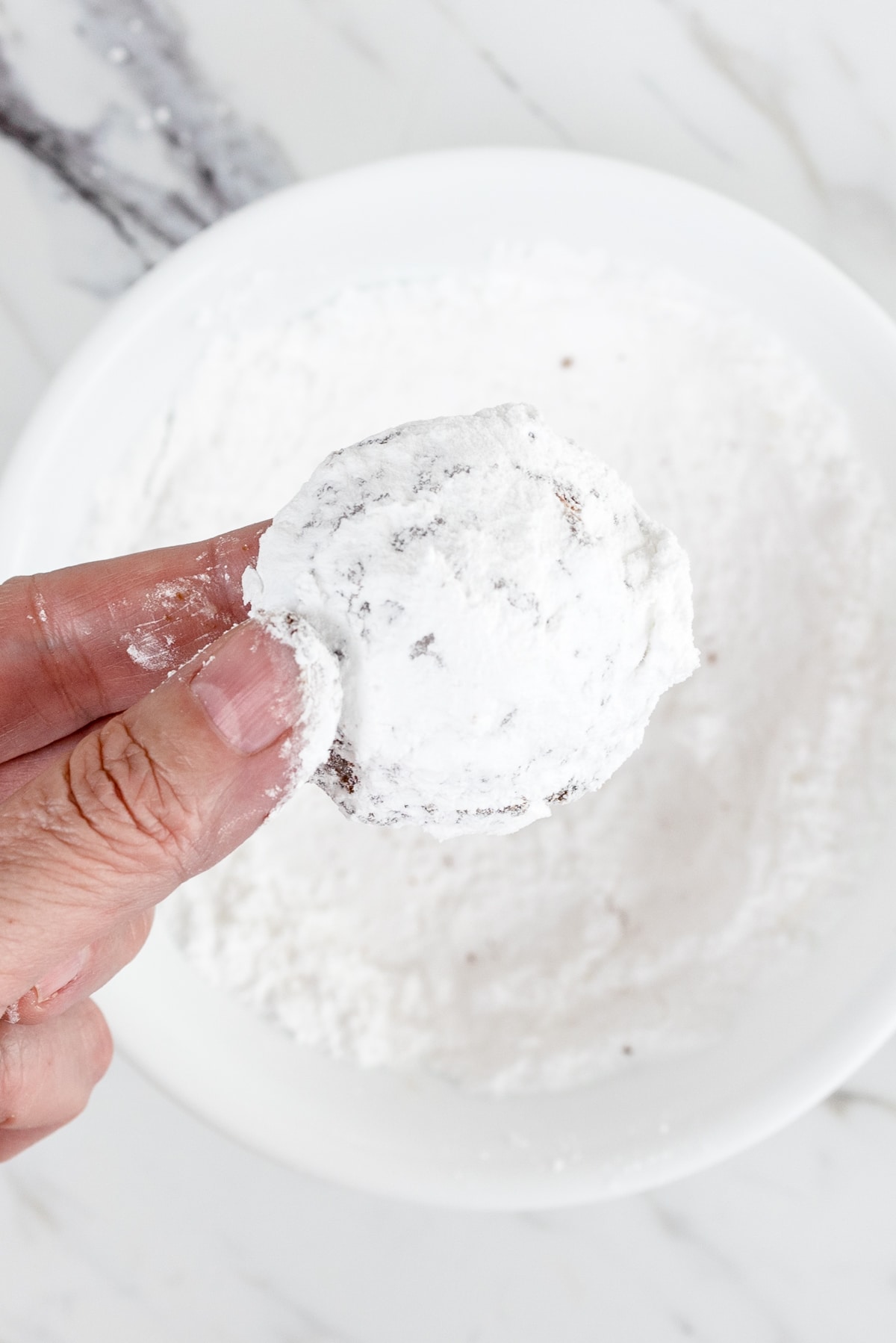 Top view of a chocolate cookie coated in powdered sugar being held above a small bowl filled with powdered sugar.