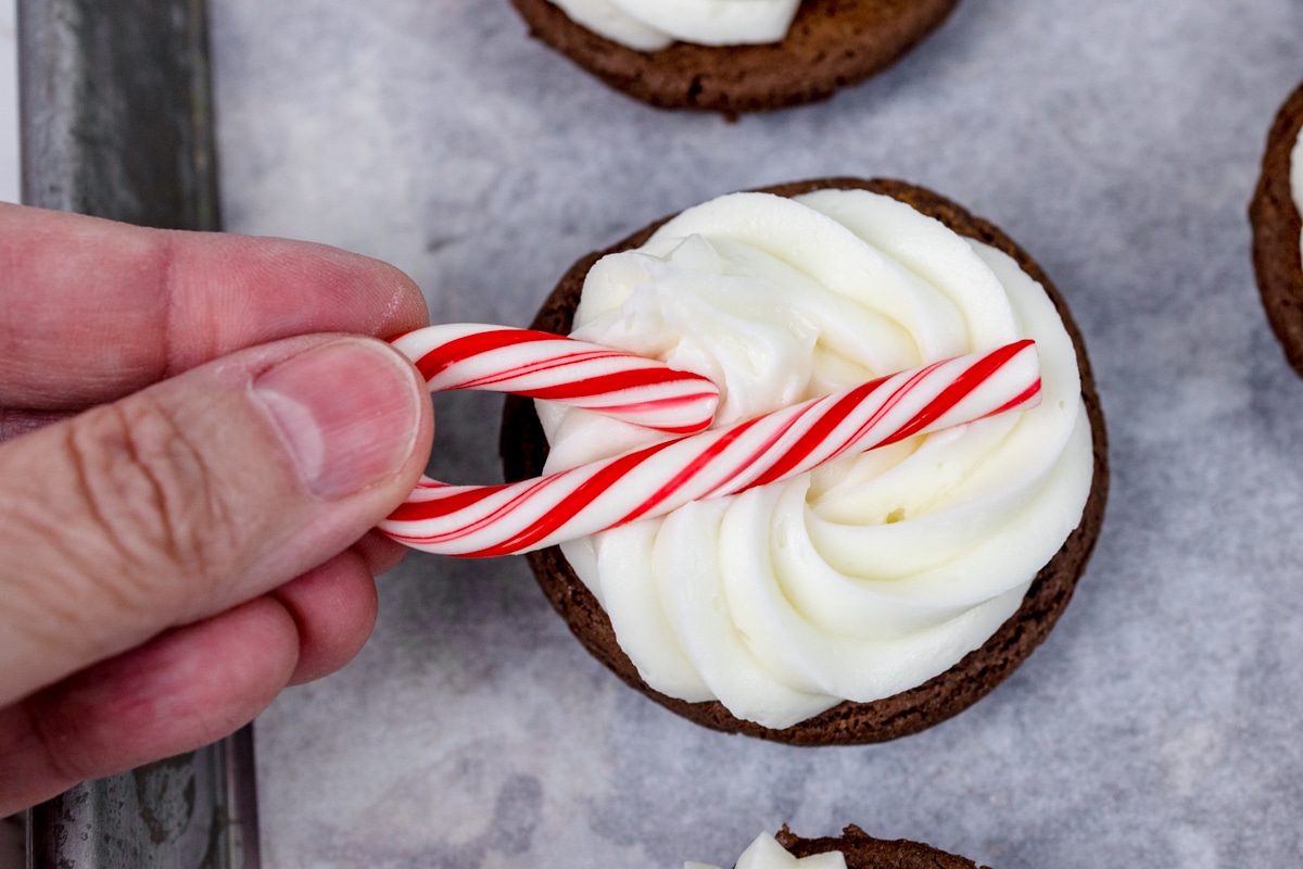 Top view of a mini candy cane being pressed into frosting on a cookie. 