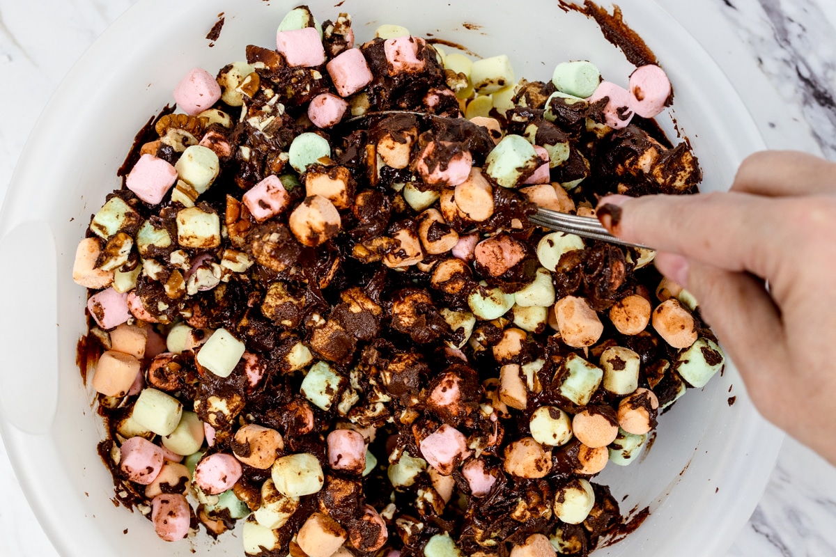 Top view of a glass mixing bowl with melted chocolate being mixed into marshmallows and nuts. 