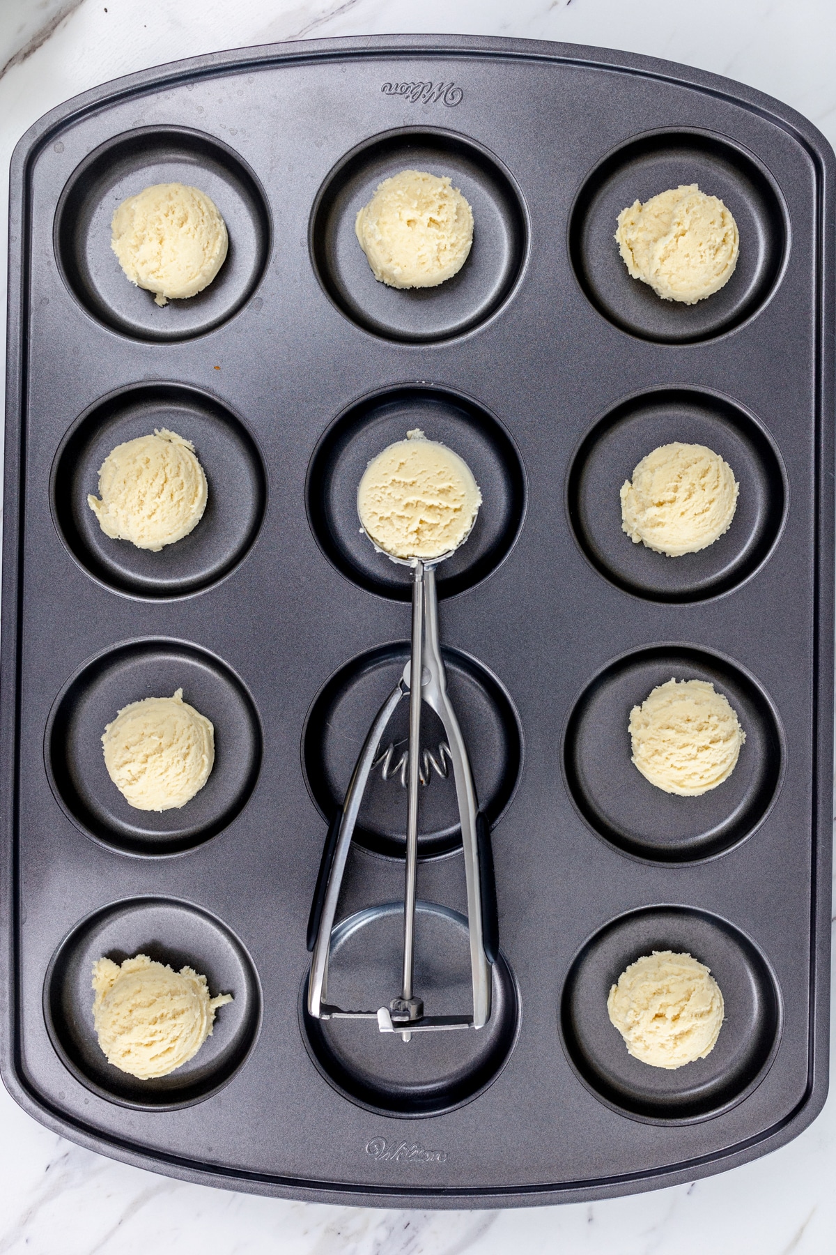 Top view of cookie dough scoops being added to a muffin top baking tray with a cookie scoop.