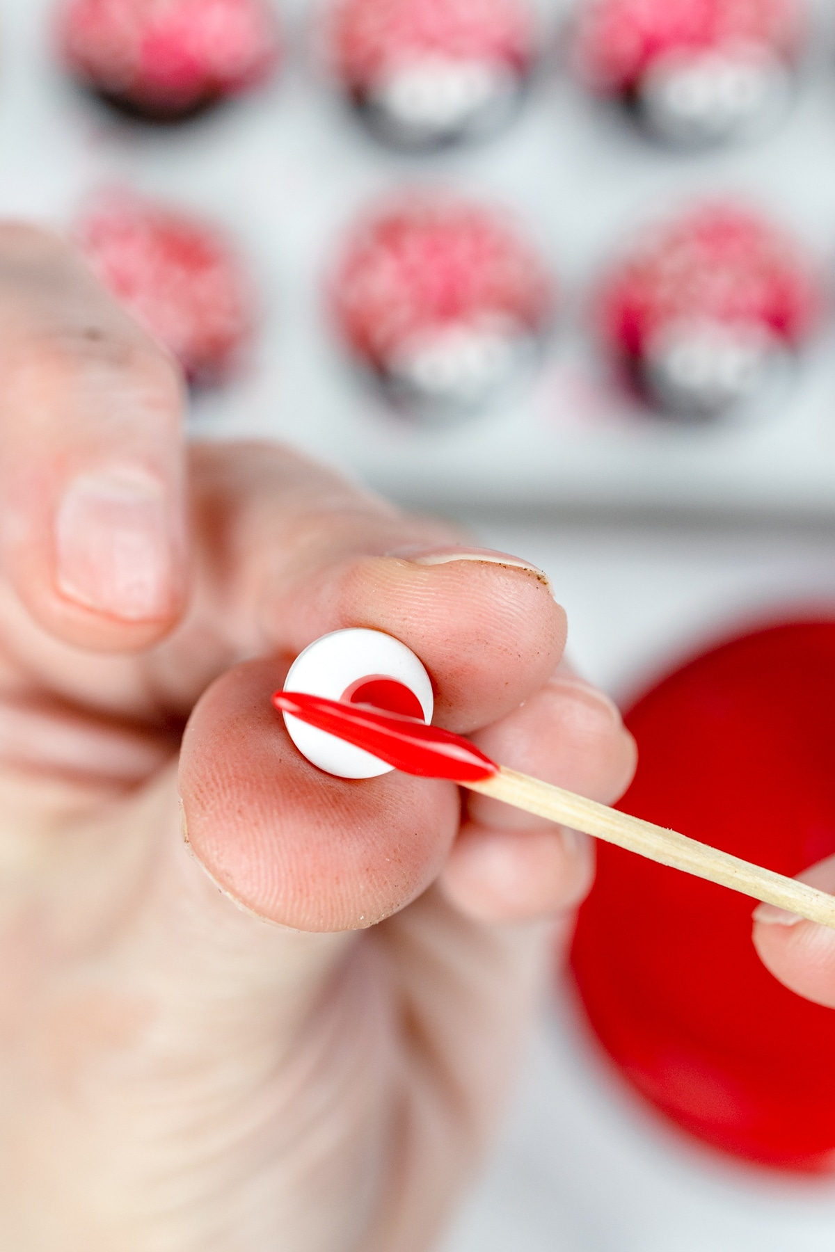 Close up of a hand holding up a candy eye ball with a toothpick being held, putting a dot of red icing onto the candy eyeball. 