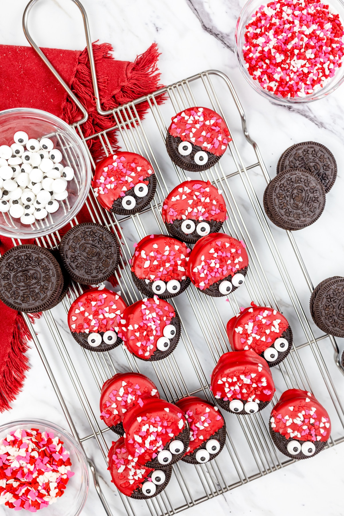 Top view of a wire rack with Love Bug Oreo Cookies scattered on it, with candy eye balls in a small bowl next to them and a small bowl of heart sprinkles next to them. 