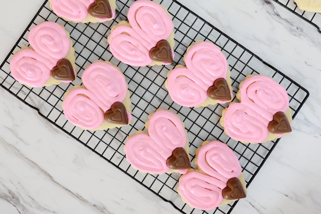 Top view of heart-shaped cookies on a wire rack with pink frosting swirled onto them to look like wings and a heart-shaped chocolate candy stuck on the tip to look like a head. 