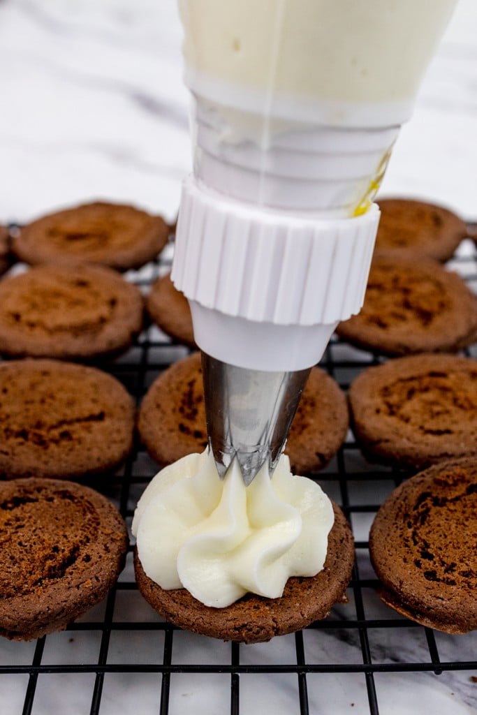 Close up of a a chocolate cookie on a wire rack being topped with frosting with a piping bag and a large tip.