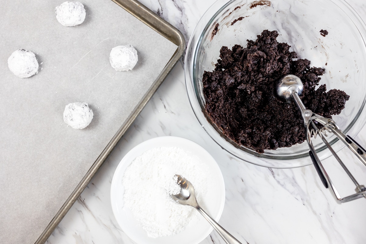 Top view of cookie mix in a glass bowl with a cookie scoop in it, next to a bowl of powdered sugar with a spoon in it, next to a baking tray filled with cookie dough balls covered in powdered sugar. 