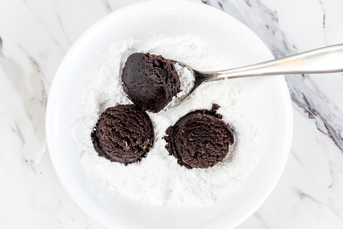 Top view of three chocolate peppermit cookie dough balls sitting in a bowl of powdered sugar with a spoon under one of them.