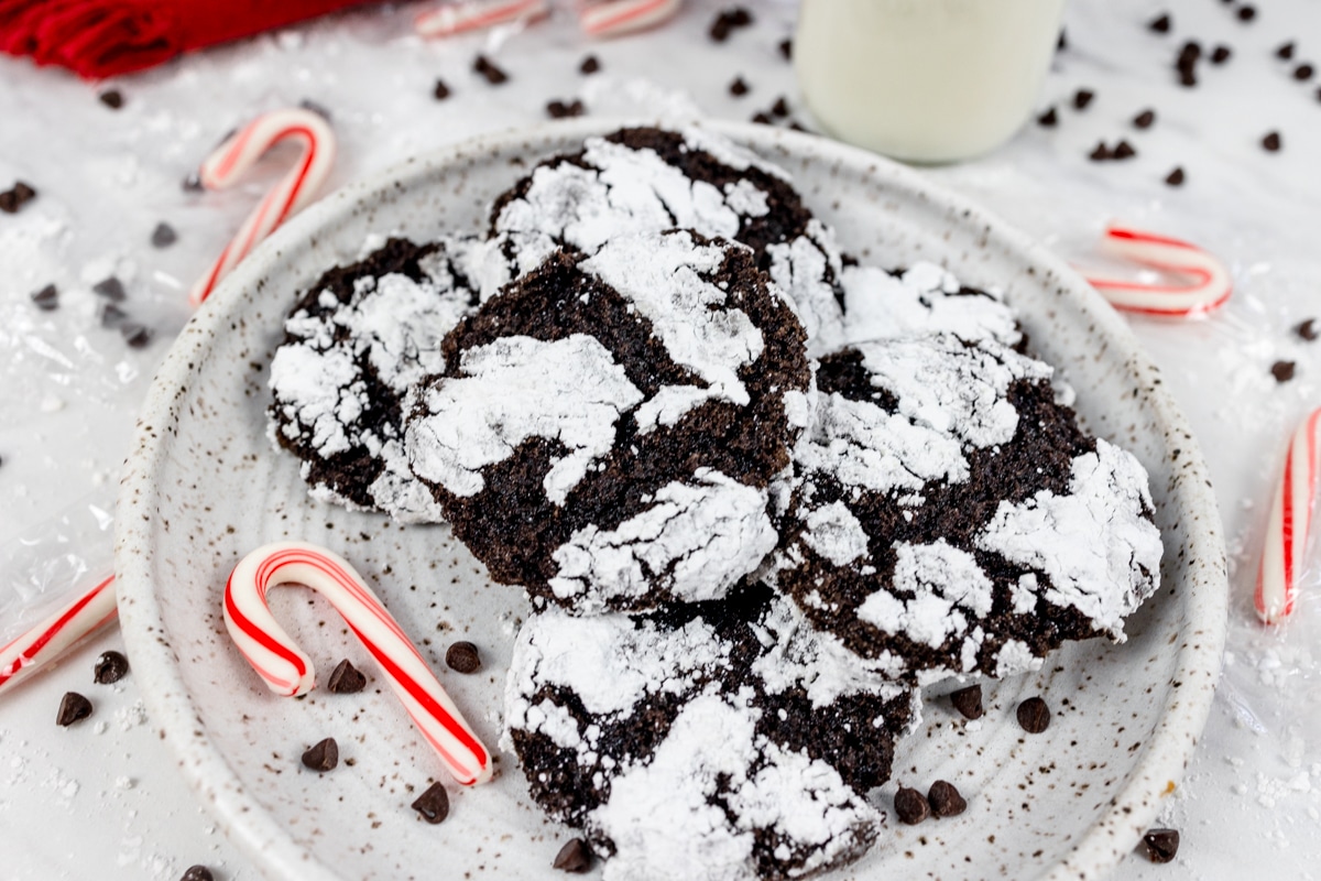 Overhead view of chocolate peppermint crinkle cookies on a white plate with a candy cane on the plate next to them. 