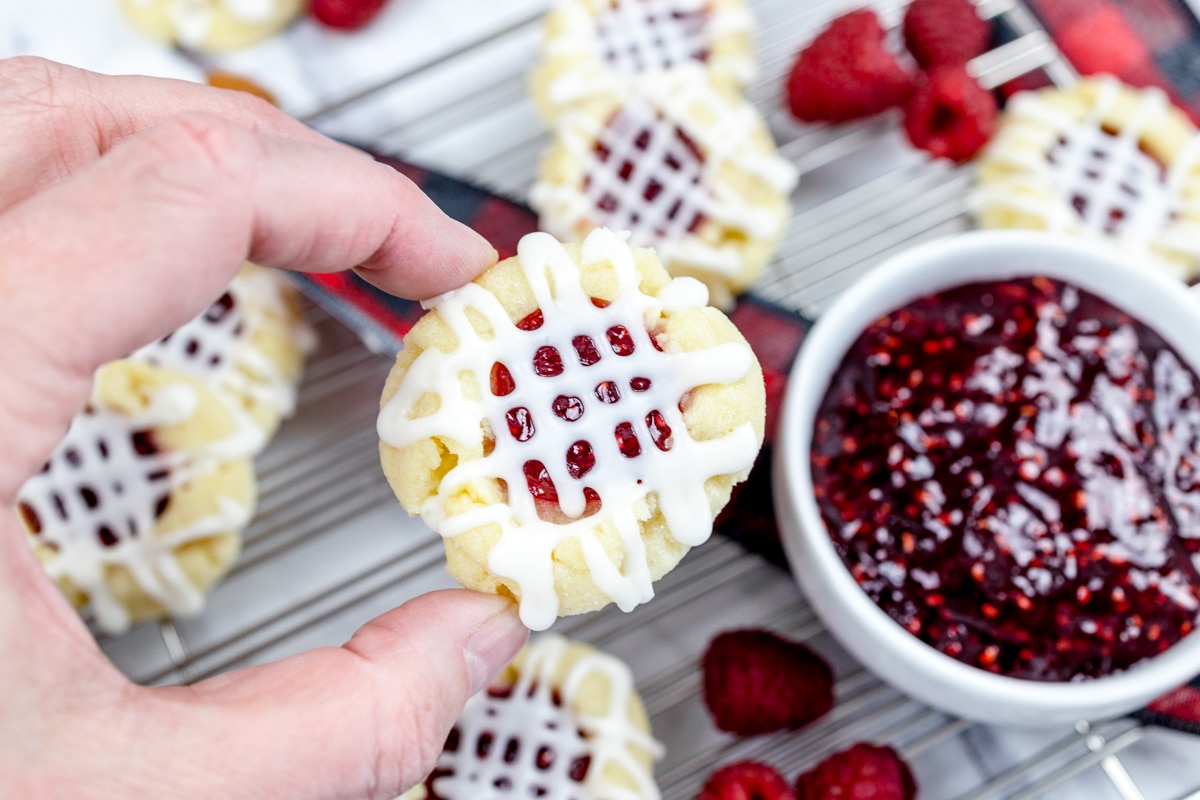 Close up of a raspberry thumbprint cookie being held in mid-air above a wire rack, next to a bowl of raspberry jam. 