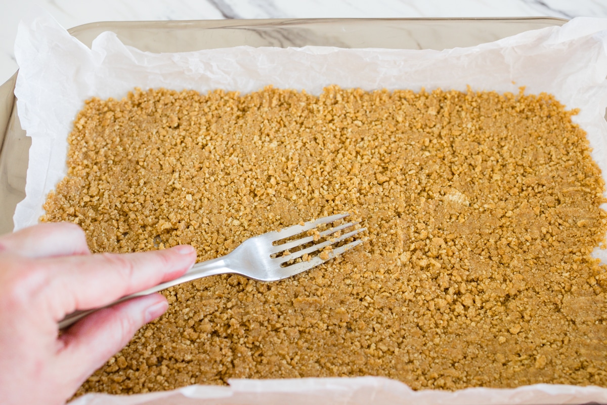 Top view of graham cracker crust being carefully patted down into an even layer at the bottom of a baking dish. 