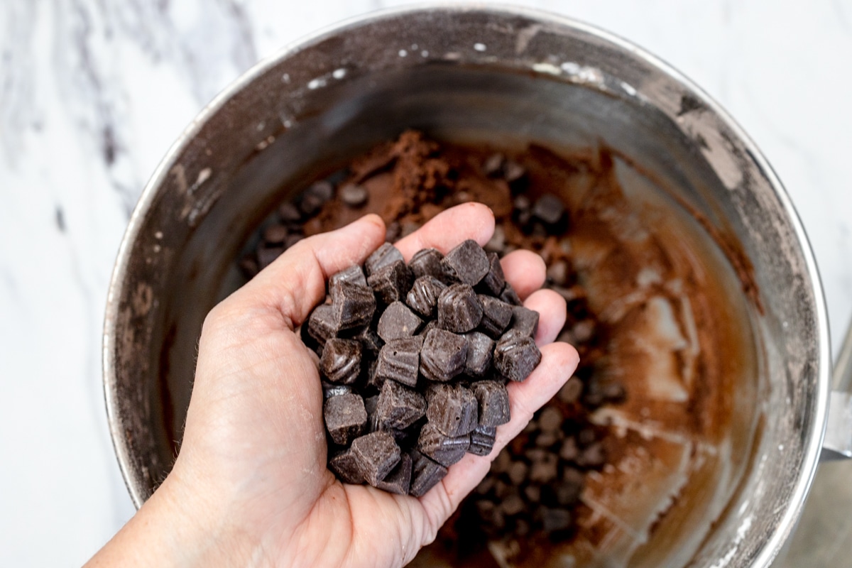Top view of chocolate chunks being added into chocolate cookie dough in a metal mixing bowl. 
