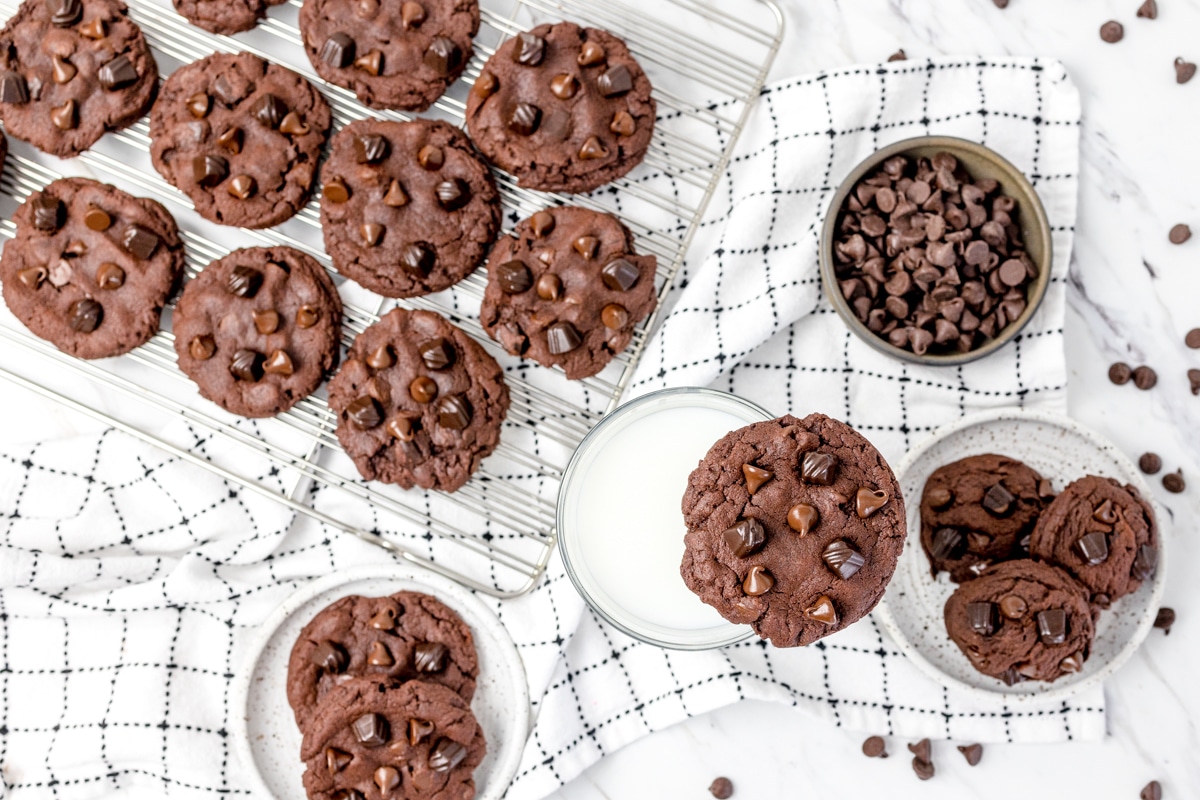 Top view of triple chocolate chunk pudding cookies on a wire rack, and on the table, with one resting on the rim of a glass filled with milk. 