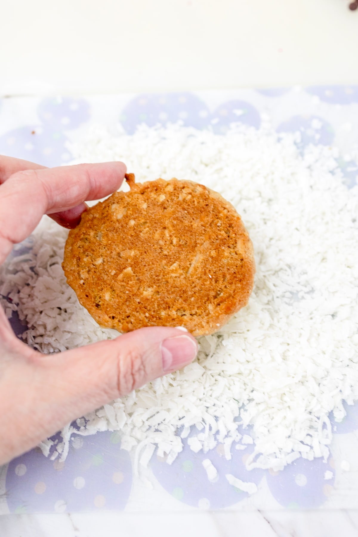 Top view of a hand dipping an oatmeal cookie into a pile pf shredded coconut. 
