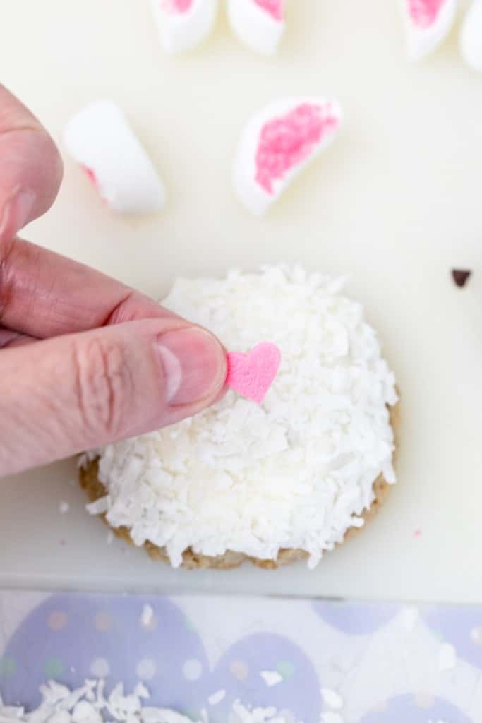 Top view of a hand putting a heart sprinkle in the center of a white cookie for a bunny nose.