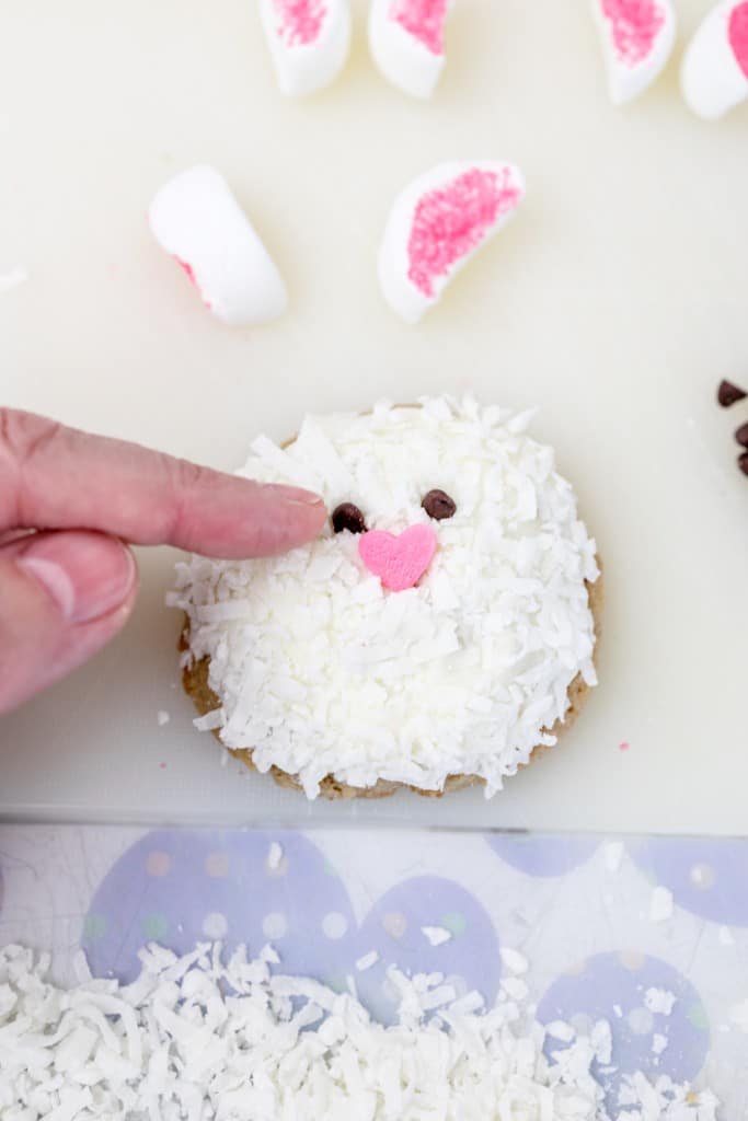 Top view of a hand putting chocolate chips above a heart sprinkle in the center of a white cookie for bunny eyes.