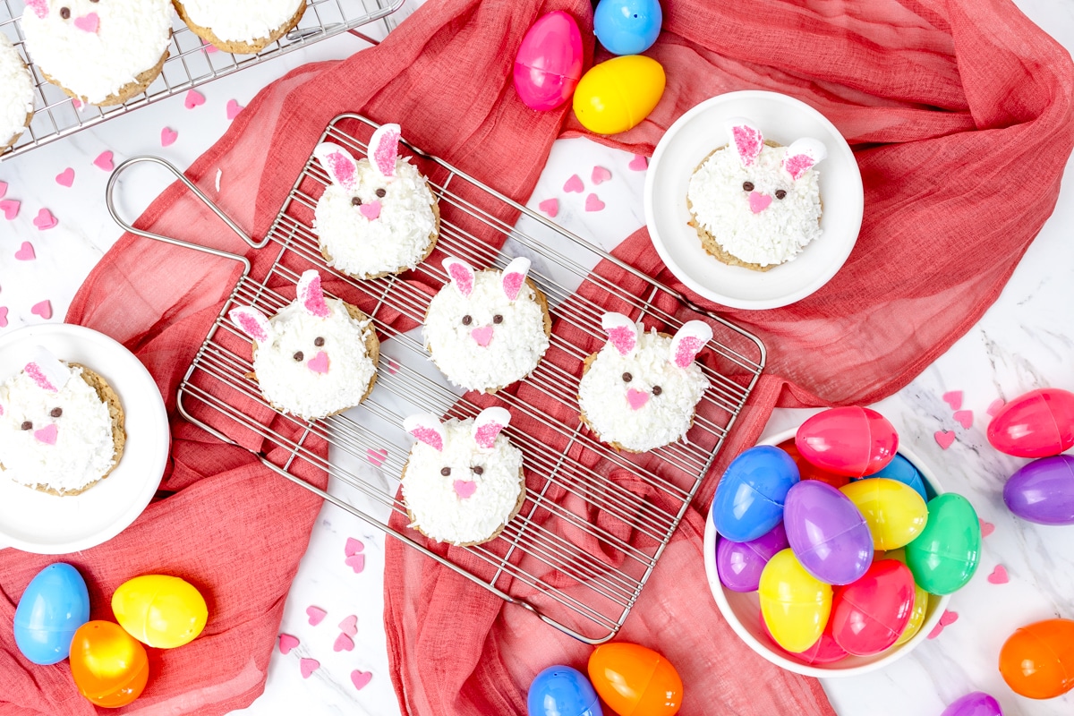Top view of Easter bunny cookies on a wire rack surrounded by colorful mini egg candies and pink heart sprinkles.