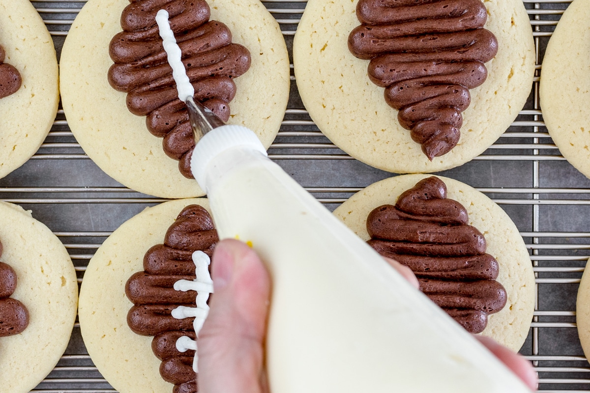Top view of round sugar cookies with brown footballs frosted onto them, and a piping bag being held above them, adding white frosting to look like stitching on the footballs. 