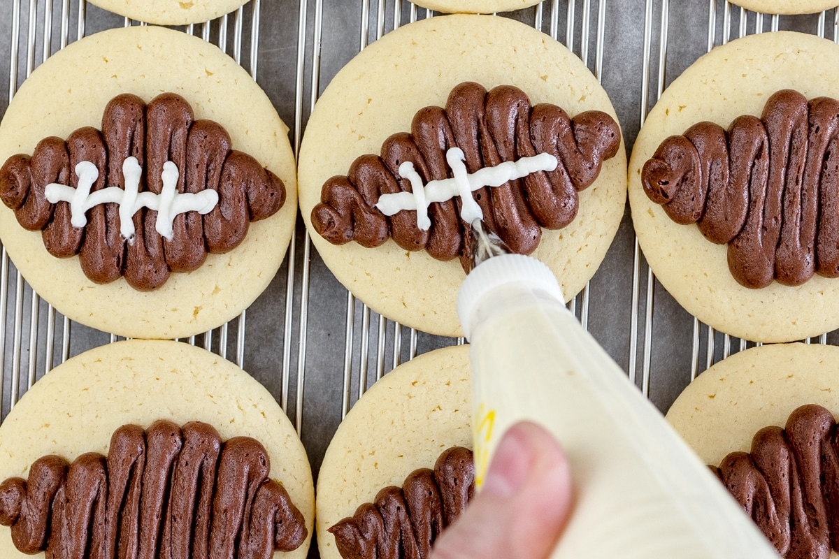 Top view of round sugar cookies with brown footballs frosted onto them, and a piping bag being held above them, adding white frosting to look like stitching on the footballs.