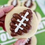 Close up of a Football Sugar Cookie in someone's hand, holding it in mid-air above a football field table backgorund.
