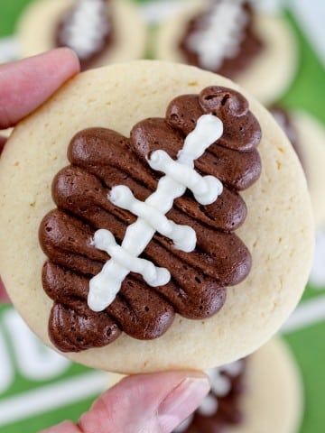 Close up of a Football Sugar Cookie in someone's hand, holding it in mid-air above a football field table backgorund.