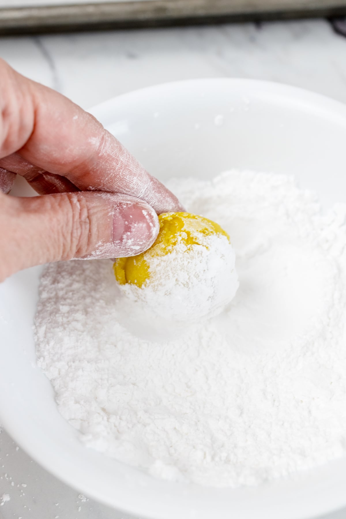 Close-up of a Lemon Crinkle Cookie dough ball being rolled in powdered sugar. 