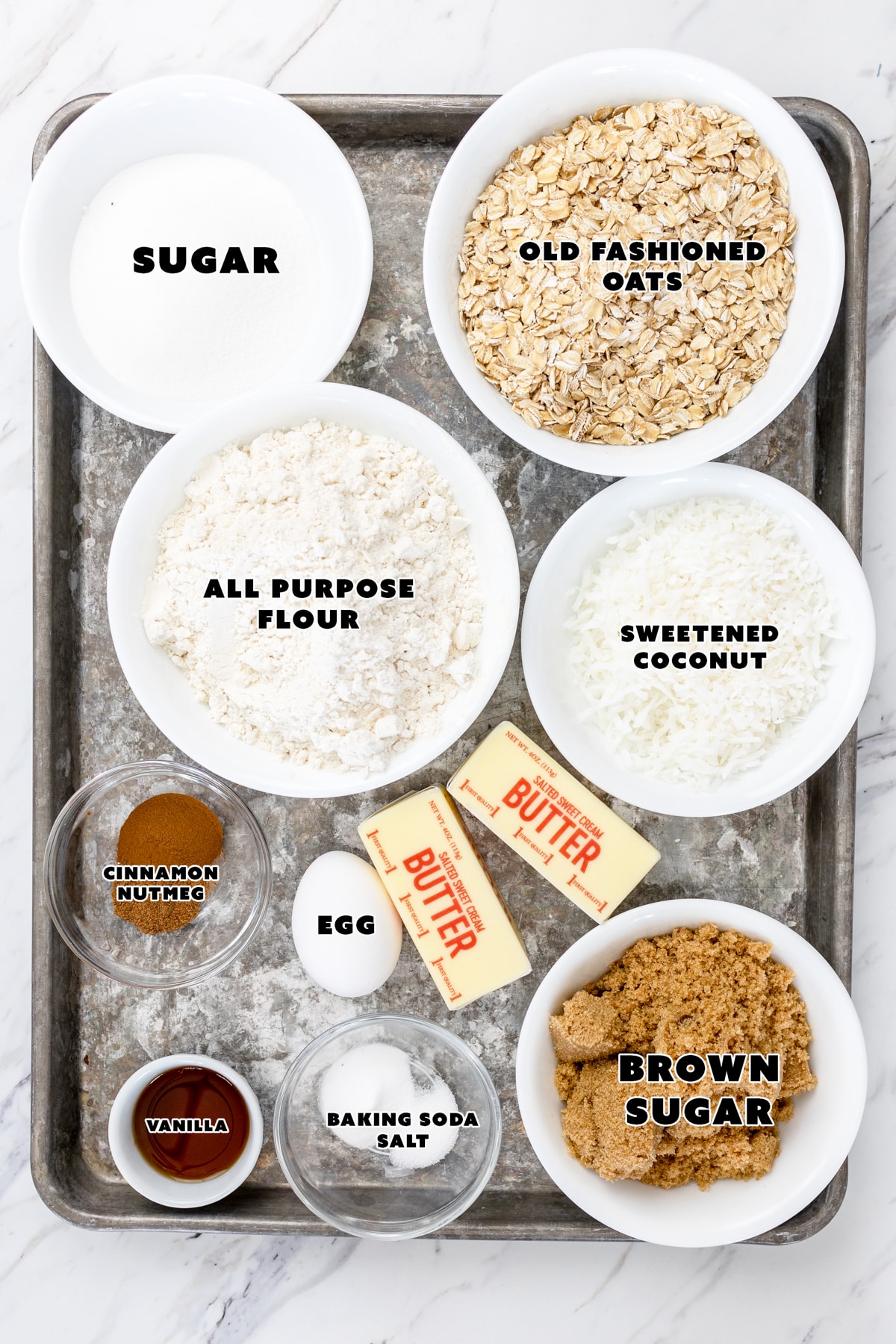 Top view of ingredients needed to make oatmeal coconut cookies, in small bowls on a baking tray. 