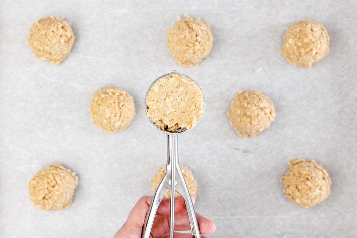 Top view of oatmeal coconut cookie dough balls on a baking tray lined with parchment paper, with one ball being held in mid-air on a cookie scoop. 