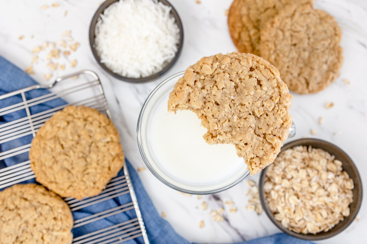 Top view of oatmeal coconut cookies on a worktop, with one oatmeal coconut cookies resting on the rim of a glass of milk. 