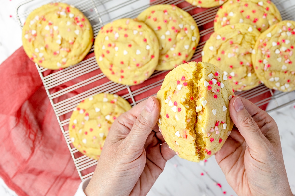 Top view of someone breaking a sprinkle sugar cookie in half above a pile of sprinkle sugar cookies in the background on a wire rack. 