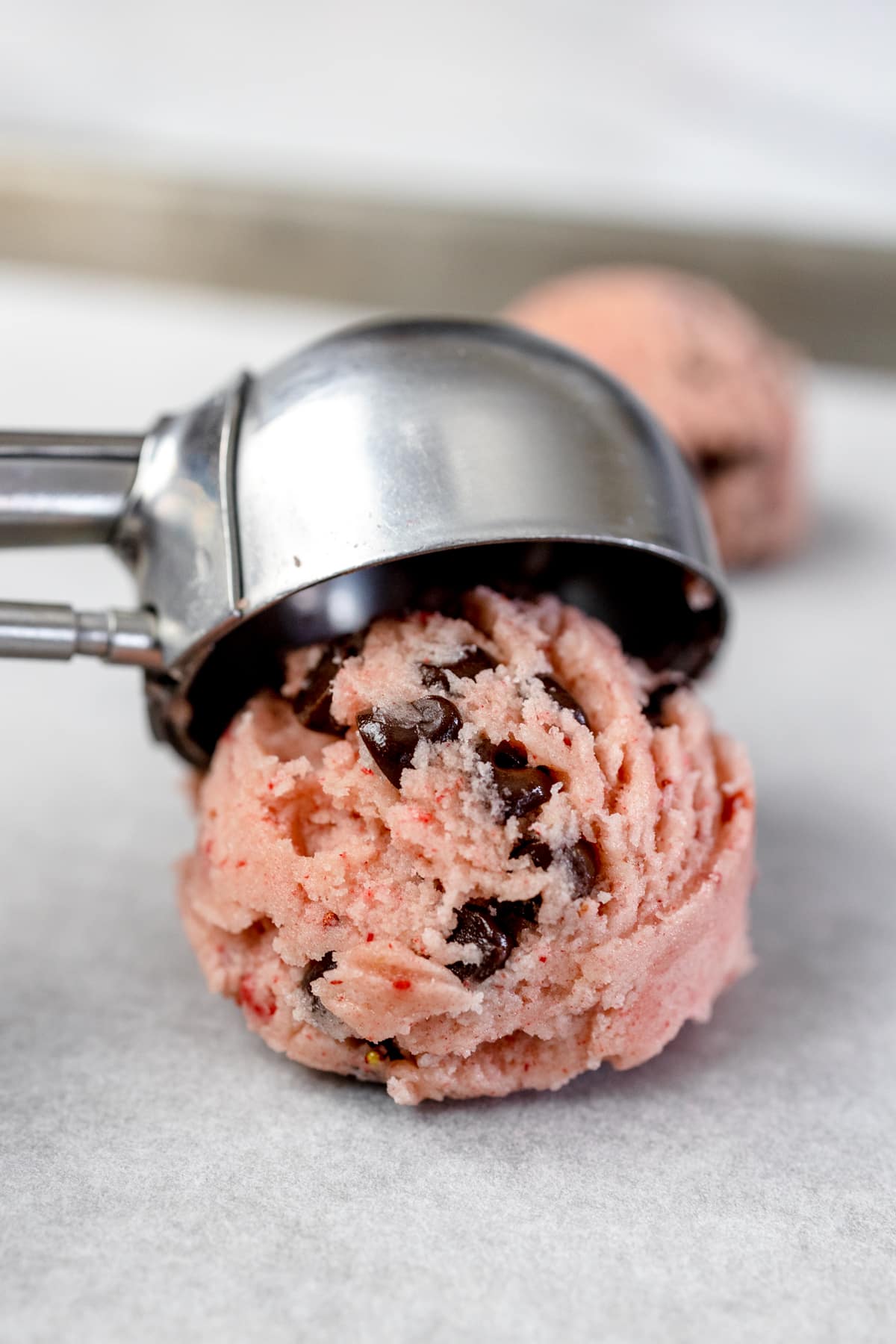 Close up of a cookie scoop putting a ball of pink chocolate chip cookie dough on a baking sheet lined with parchment paper. 
