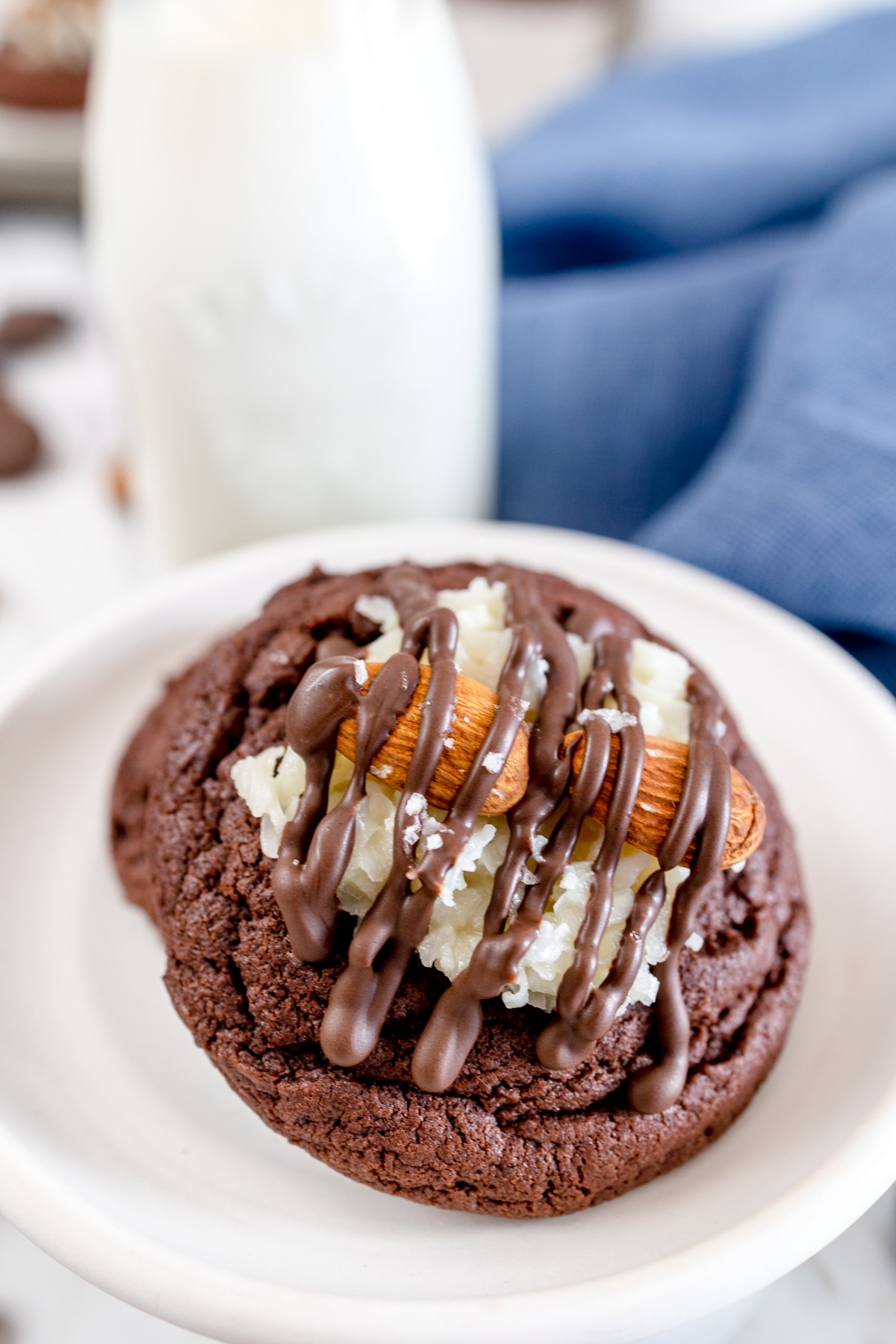 Close up of an Almond Joy Cookie on a white plate with a glass bottle of milk behind it. 